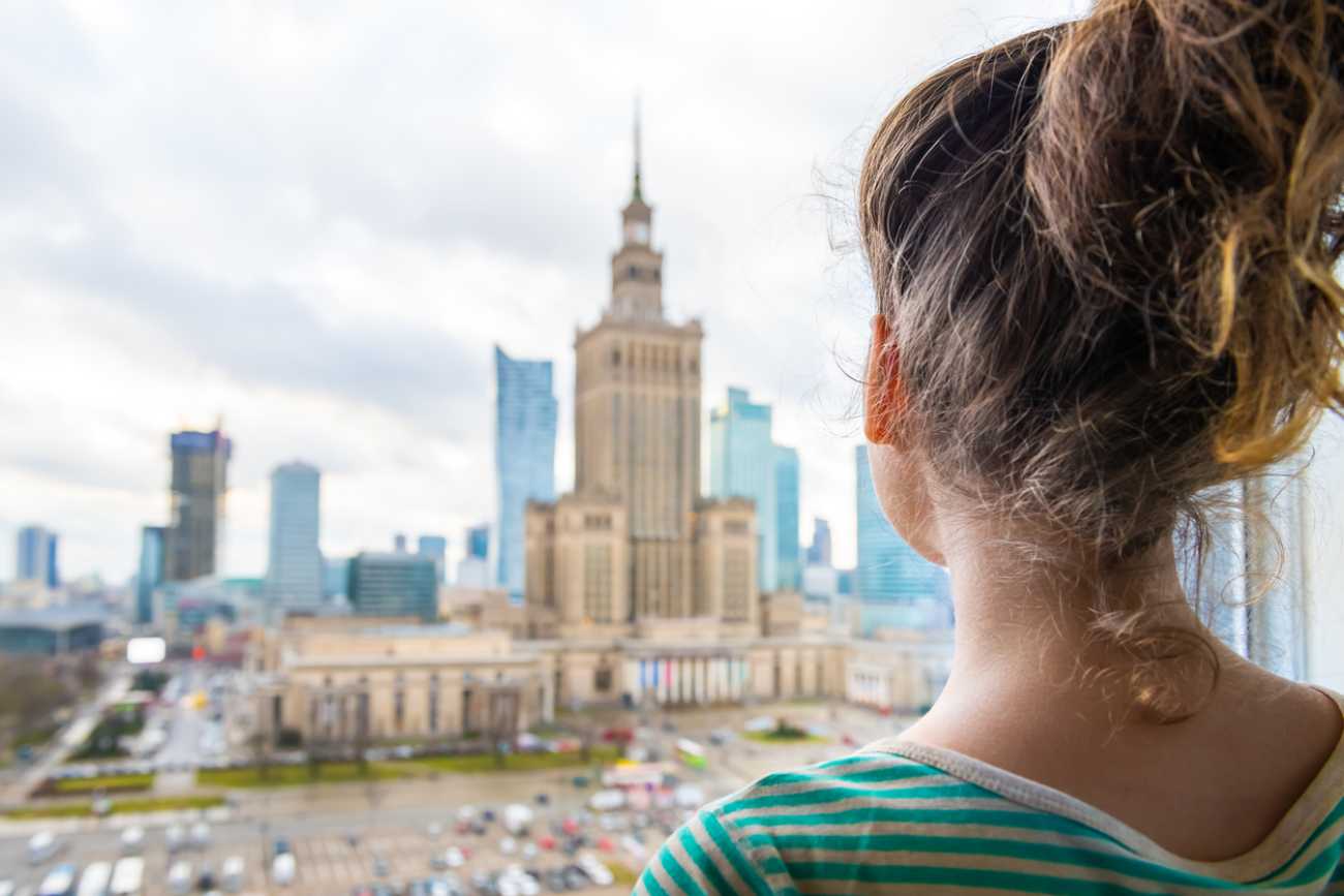 Kobieta stojąca przy oknie w mieszkaniu patrzy na Pałac Kultury i Nauki w Warszawie widoczny w tle za szybą.